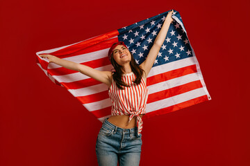 Happy young hipster woman carrying American flag while standing on red background