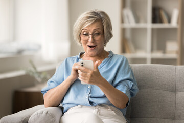 Surprised excited senior woman using mobile phone on home couch, staring at smartphone screen,...