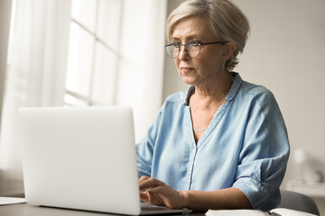 Focused old mature business woman in elegant glasses typing on laptop computer, working at home, using modern Internet technology for job communication