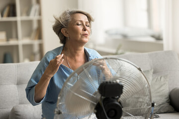Exhauster older retired woman sitting at electric fan with propeller, enjoying cool fresh air flow, feeling relief, sitting on home sofa, suffering from hot weather, overheating - obrazy, fototapety, plakaty