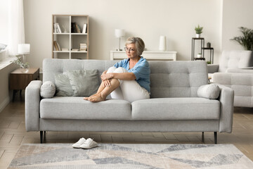 Depressed older retired woman sitting on sofa at cozy modern home, looking away in deep thoughts, staring into vacancy, thinking on depression, apathy, loneliness, remembering - obrazy, fototapety, plakaty