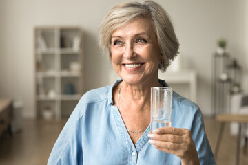 Happy satisfied elderly lady holding transparent glass pf pure clean water, standing in home...