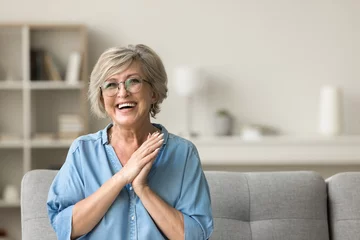 Photo sur Plexiglas Vielles portes Cheerful pretty older woman in elegant glasses sitting on cozy home couch, smiling with perfect white teeth, laughing with hands at chest gesture, enjoying leisure, comfort, having fun