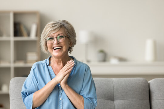 Cheerful pretty older woman in elegant glasses sitting on cozy home couch, smiling with perfect white teeth, laughing with hands at chest gesture, enjoying leisure, comfort, having fun