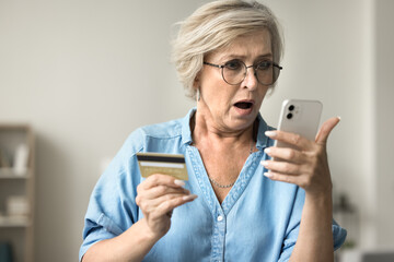 Shocked concerned mature woman in glasses holding credit card, staring at smartphone in bad surprise, getting stress, financial problems, money stealing, overspending, bankruptcy risk - obrazy, fototapety, plakaty