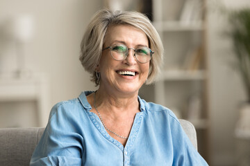 Happy positive blonde elder woman in stylish eyeglasses looking at camera with perfect toothy smile, laughing, posing for home portrait. Beautiful mature lady video call screen head shot