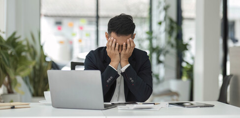 Young unhappy man office worker feeling bored at work, looking at laptop with demotivated face expression while sitting at workplace in office, distracted male worker feeling tired of monotonous job