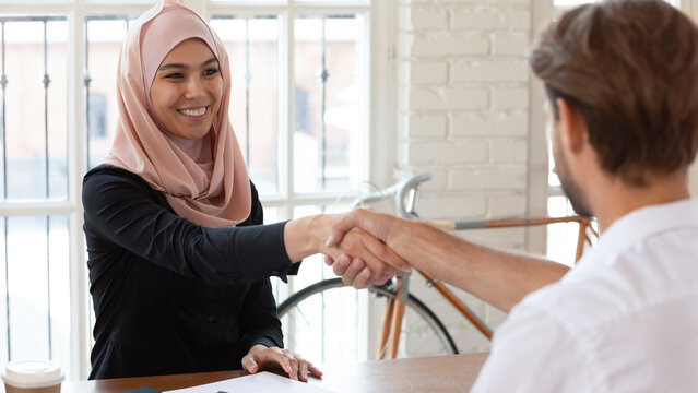 Seated at office desk smiling middle eastern ethnicity businesswoman shakes hand european businessman greeting each other starting business meeting, makes offer financial deal, express appreciation