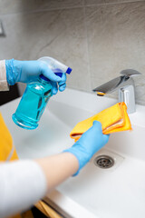 Female hands wearing rubber gloves holding microfiber cloth and spray bottle sanitizer to disinfect sink tap faucet in bathroom.