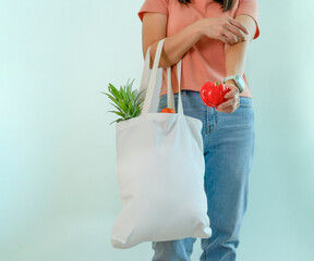 a female one hand holds red bell pepper,other hand carry reusable cloth bag of fruits and vegetables isolated on blue background,concept campaign of using reusable bag,zero waste, stop global warming