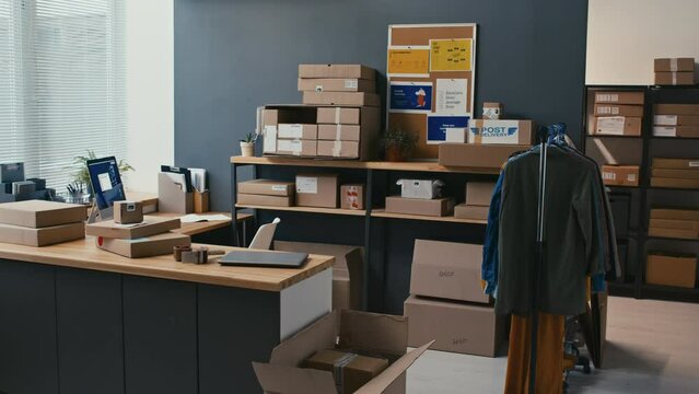 Long shot of spacious storage room with boxes on shelves, rack with second hand clothes and writing table with laptop
