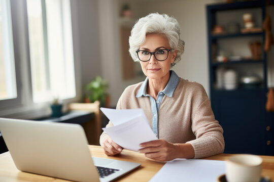 Adult senior 60s woman working at home at laptop.  middle aged woman at table holding document calculating bank loan payments, taxes, fees, and retirement finances. Copy space. Website images