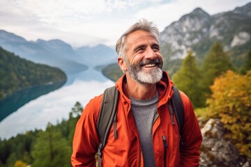 Naklejka na ściany i meble Amidst the rugged beauty of nature, a man in an orange jacket stands by the tranquil river, a smile on his face as he takes in the vast expanse of sky, water, and mountains surrounding him