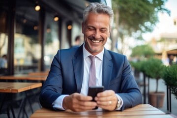 A sharply-dressed businessman exudes confidence as he flashes a charming grin while scrolling through his phone at an outdoor table, his tailored suit and tie complementing the sleek furniture - obrazy, fototapety, plakaty