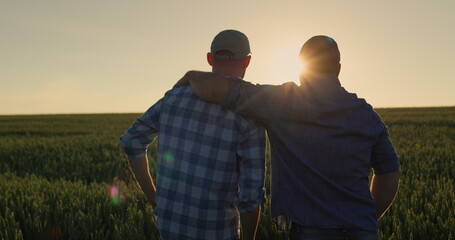 The farmer hugs his adult son and looks forward together to the wheat field where the sun is...