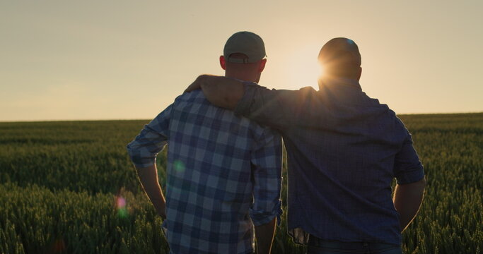 The farmer hugs his adult son and looks forward together to the wheat field where the sun is setting. Family business and traditions concept