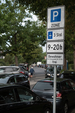 Schild Parkzone "Anwohnerparken" vor zugeparkten Wegen in der N&auml;he des Hamburger Flughafens. Alte Frau mit Gehstock auf Fahrbahn neben SUV.