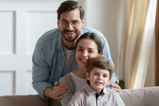 Head shot portrait happy young family with cute little son, pretty preschool boy sitting on beautiful mother laps, smiling husband wearing glasses touching wife shoulders, posing for photo