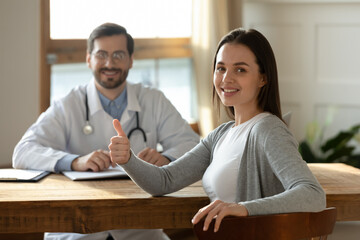 Smiling young woman patient showing thumb up gesture at medical appointment, sitting at desk in friendly male doctor office in hospital, satisfied client recommending healthcare service, good quality