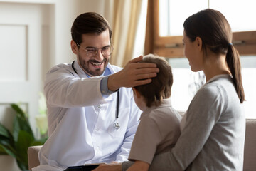 Smiling caring doctor wearing glasses and white uniform stroking little patient head, pediatrician showing caress and support, cute preschool boy sitting on mother laps during medical appointment