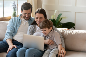 Happy young parents with cute little son using laptop together, looking at screen, smiling mother and father wearing glasses with preschool child watching cartoons, shopping online, sitting on couch