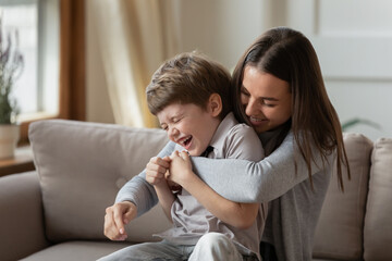 Happy young mother and little son having fun on cozy couch in living room, overjoyed mum and preschool child tickling, cuddling, enjoying leisure time together, playing funny active game at home