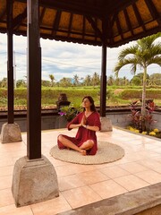 Young beautiful woman meditating and doing yoga in red dress in abandoned temple with green palms and rise field background on Bali. Female practicing pose. High quality FullHD vertical footage