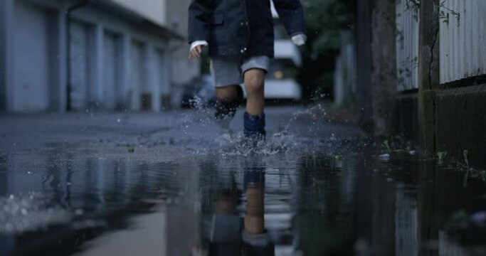 Carefree child running with rainboots on water puddles in street during evening in ultra slow-motion 800 fps splashing droplets in the air
