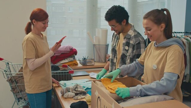 Medium shot of female coordinator of charity project shooting video on camera of her smartphone of volunteers preparing clothing to ship to poor people