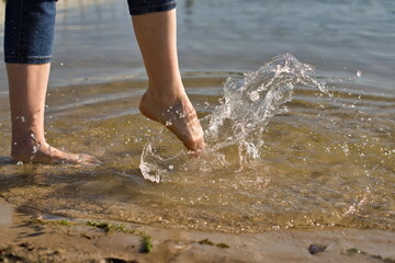 Noga osoby w spodniach kopiącej wodę na brzegu jeziora i rozbryzg kropelek w słoneczny letni dzień - obrazy, fototapety, plakaty