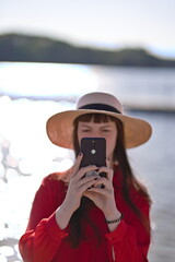 Kobieta w kapeluszu i czerwonej koszuli robiąca zdjęcie telefonem dla fotografa na tle jeziora w słoneczny letni dzień - obrazy, fototapety, plakaty