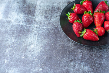 Red, ripe, juicy strawberries in a black plate on a light stone table.