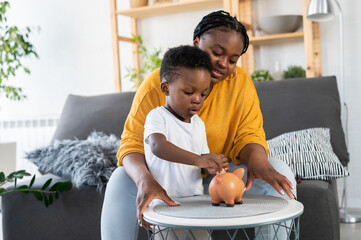 Cute little African-American boy with his mother inserting a coin into a piggy bank, financial and banking concept. Child saving money for the future concept.