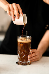 Barista woman pouring coffee on ice cubess in high old fashioned glass with tonic served on table in coffee shop