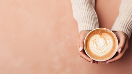 Top view of a female hands in a warm sweater holding mug of delicious cappuccino coffee. Pastel pink background with copy space, banner template.  - obrazy, fototapety, plakaty