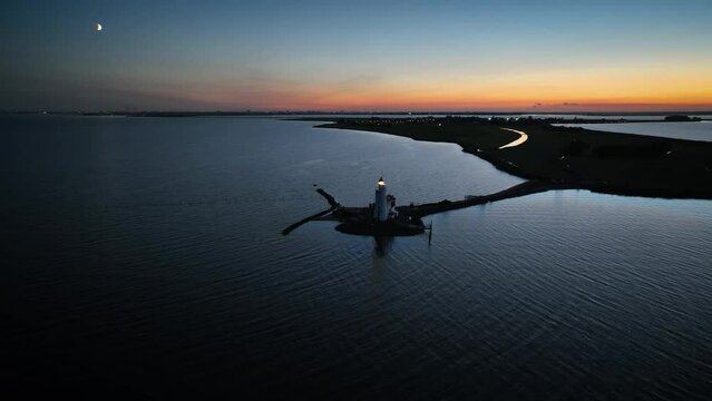 Circling around lighthouse Paard van Marken in Nethelands at dawn. Aerial footage of Dutch peninsula Marken by evening sun, on the IJsselmeer. The lighthouse's beam rotates, casting its guiding light.