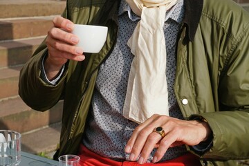 Sitzender Mann mit grauem Hemd mit Muster, weißem Schal und grüner Jacke hält weiße Tasse...