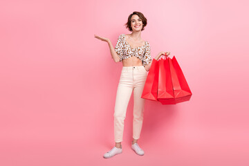 Full length photo of pretty woman wear print top white pants hold new outfit palm presenting empty space isolated on pink color background