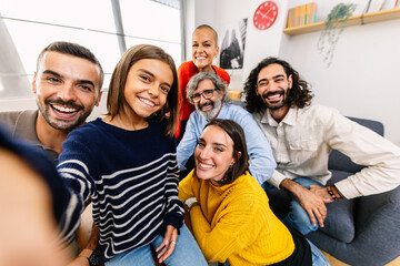 Happy self portrait of multi-generational family at home. Little cute girl taking selfie with parents, uncle, aunt and grandfather sitting together on sofa. Mobile phone screen on video call.