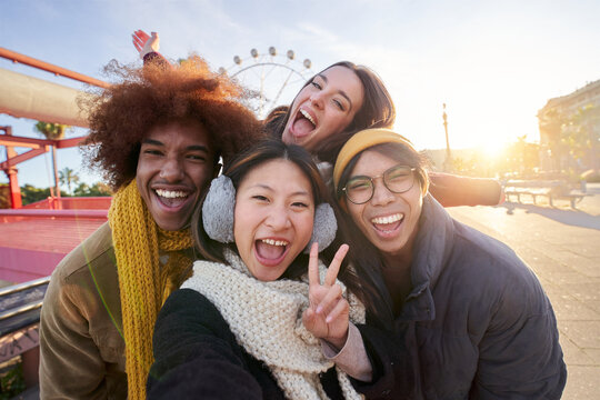 Excited smiling group of multiracial friends taking funny selfie phone looking at camera standing together outdoor. Multi-ethnic young people in amusement park having fun in sunny winter holiday. 