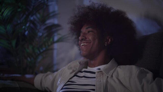 African American cheerful man afro hair sitting happy on sofa laughing excited. Young casual generation z male smile enjoying funny comedy movie or sitcom series and turn on TV indoors home at night. 