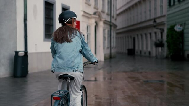 Back view of beautiful young woman riding bicycle along the rainy street at city center