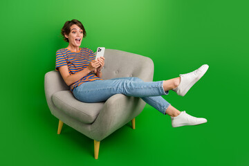 Full body photo of astonished positive lady sit comfort chair use smart phone isolated on green...