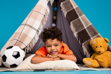 Photo of cute boy kid having nap in diy tent playing with toys lying isolated blue color background © deagreez