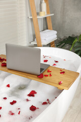 Fototapeta na wymiar Wooden board with laptop and rose petals on bath tub in bathroom