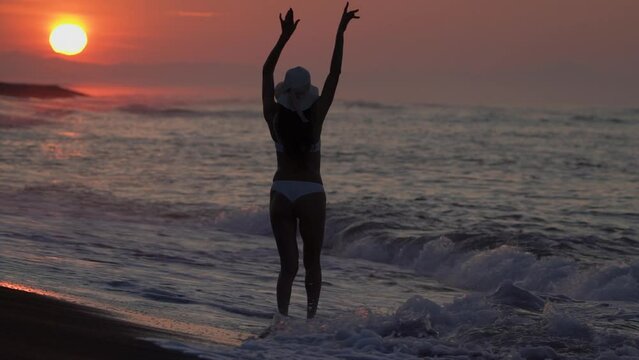 Rear view of unrecognizable woman in white bikini standing on beach ankle deep in water breaking waves of ocean at sunrise on summer morning during beach holiday. Slow motion, cinematic shot, handheld