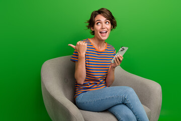 Photo of astonished charming girl sit chair hold smart phone look indicate finger empty space isolated on green color background