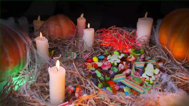 Happy Halloween scary background with pile colorful sweets and creepy bone hand with candy slowly reaches out, burning candles, pumpkins in cobweb among dry grass in darkness. Spirits night.