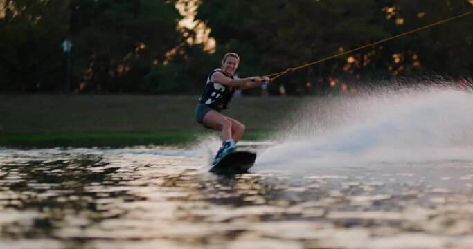 Female wakeboarder wakesurfing at sunset. Girl riding on waterski cabel. Holding tow rope. Recorded at 50fps on cinema camera.