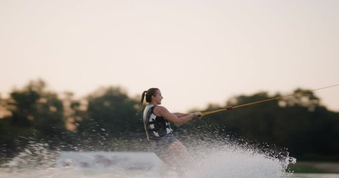 Female wakeboarder wakesurfing at sunset. Girl riding on waterski cabel. Holding tow rope. Summer activities in a lake. 120fps cinema camera.